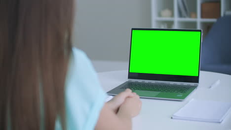 Woman-sits-at-desk-in-bedroom-she-looks-at-laptop-green-screen-and-talks-to-someone-over-internet-video-communications-sometimes-taking-notes-in-notebook.-Close-up